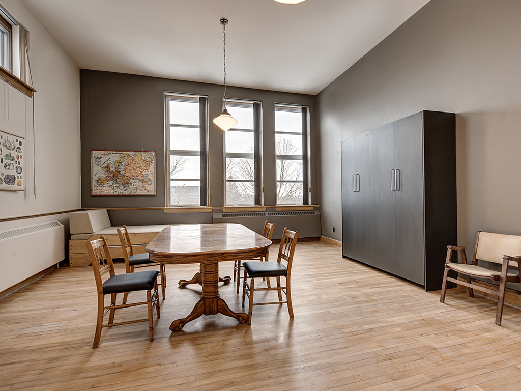 Coin repas avec table en bois, chaises et grandes fenêtres à l'Espace du Carmel